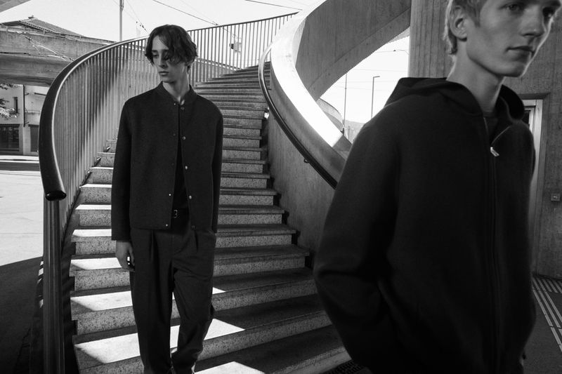 Two adult male models pose on concrete steps outdoors, one in jacket and pants, the other in hoodie, in a fashion editorial image.