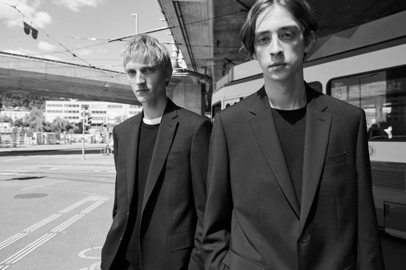 Two young adult men in dark suits pose outside near a light rail or streetcar line in a black and white editorial fashion image.