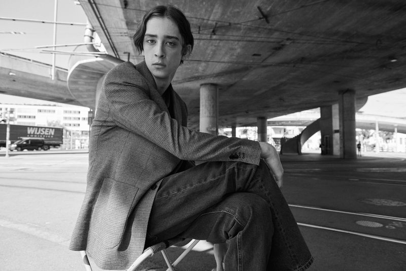 Black-and-white fashion shot of a young adult male in a suit jacket and jeans, seated outdoors under a concrete structure on a sunny day.