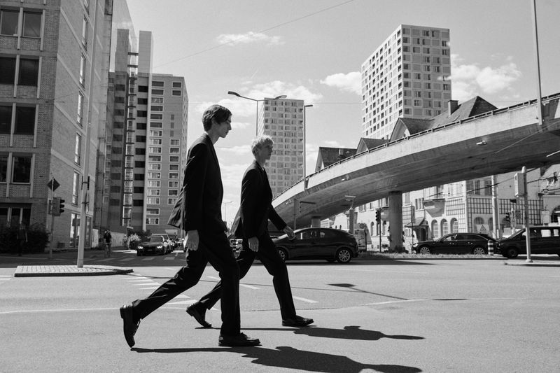 Two men in black suits walking across a city street on a sunny day, featuring modern architecture and crossing path.