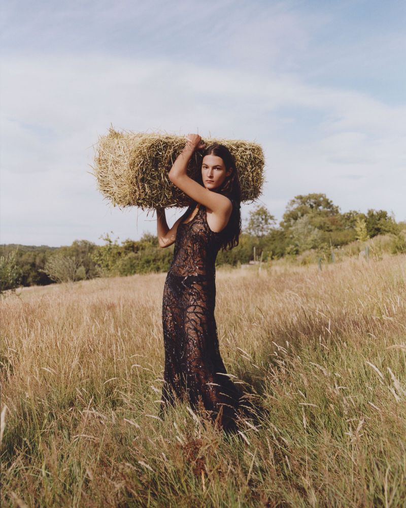 Woman in an elegant black lace dress balances a hay bale on her shoulders in a golden field on a sunny day, exuding rural fashion.
