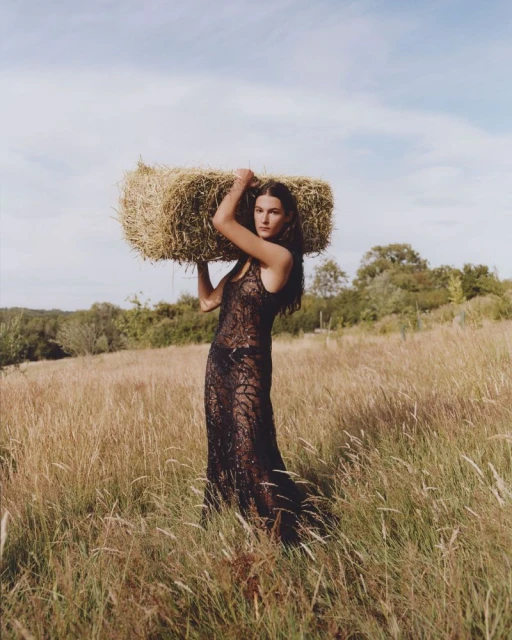 Woman in an elegant black lace dress balances a hay bale on her shoulders in a golden field on a sunny day, exuding rural fashion.