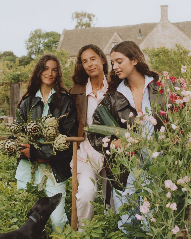 Three women in a garden surrounded by lush green plants and flowers, holding fresh produce and flowers, with a black dog nearby.
