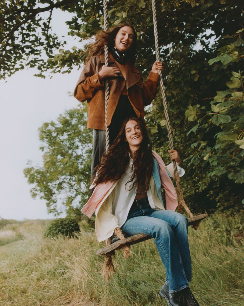 Two smiling women on a rope swing in a grassy, wooded area; one wears a brown jacket and the other a colorful shirt, set in daylight.