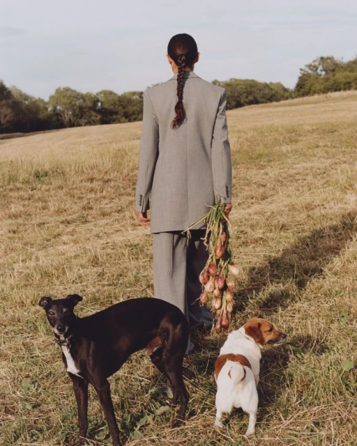 Woman in suit with braided hair stands in field, carrying onions. Two dogs are in the foreground, one black and one white.