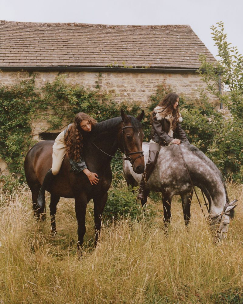 Two young women riding horses in a grassy field with a stone building in the background. Equestrian lifestyle in rural setting.