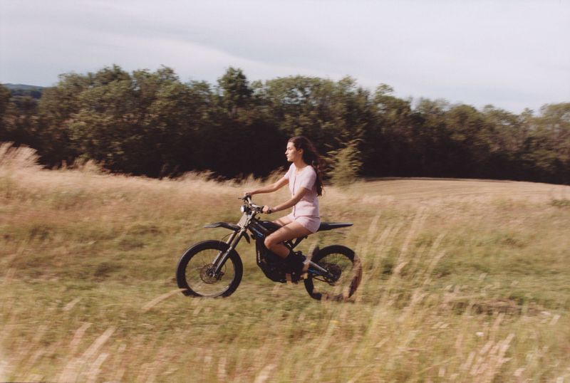 Young woman in pink outfit riding a black dirt bike through a grassy field on a sunny day with trees in the background.