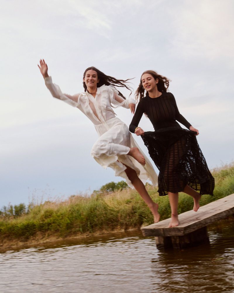 Two young women in long dresses leap barefoot from a wooden platform towards a river in a grassy, rural landscape under a cloudy sky.