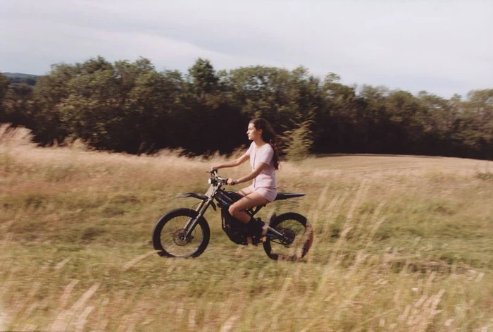 Woman riding a dirt bike through a grassy field with trees in the background under an overcast sky.