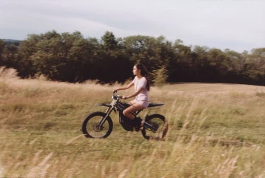 Woman riding a dirt bike through a grassy field with trees in the background under an overcast sky.