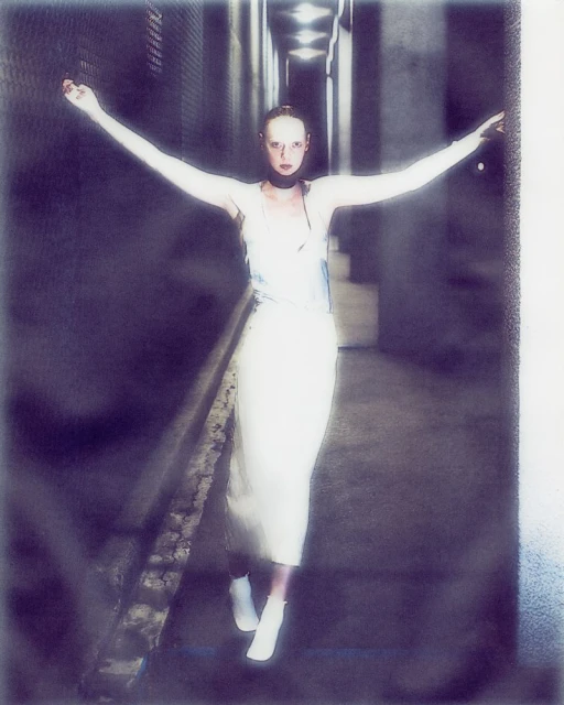 Woman in a long white dress stands in a gray hallway with arms outstretched, lit by overhead lighting and wearing white ankle boots.