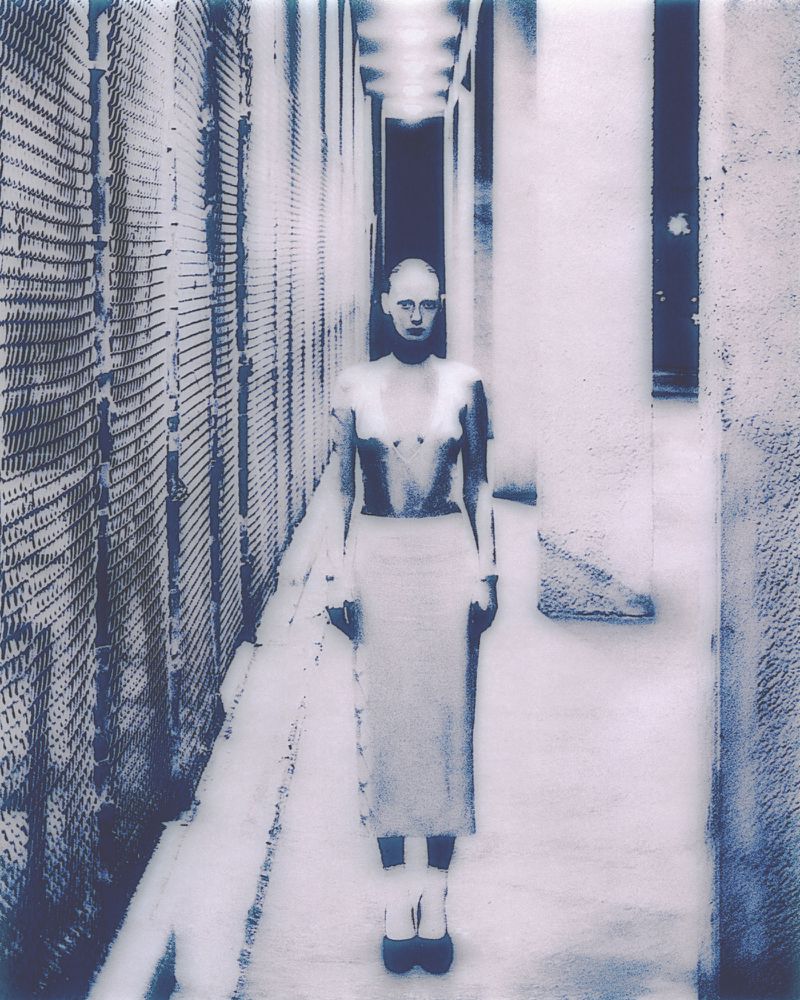 Monochromatic art image of a slim woman with a shaved head standing in a bright, narrow hallway with light shining from above.