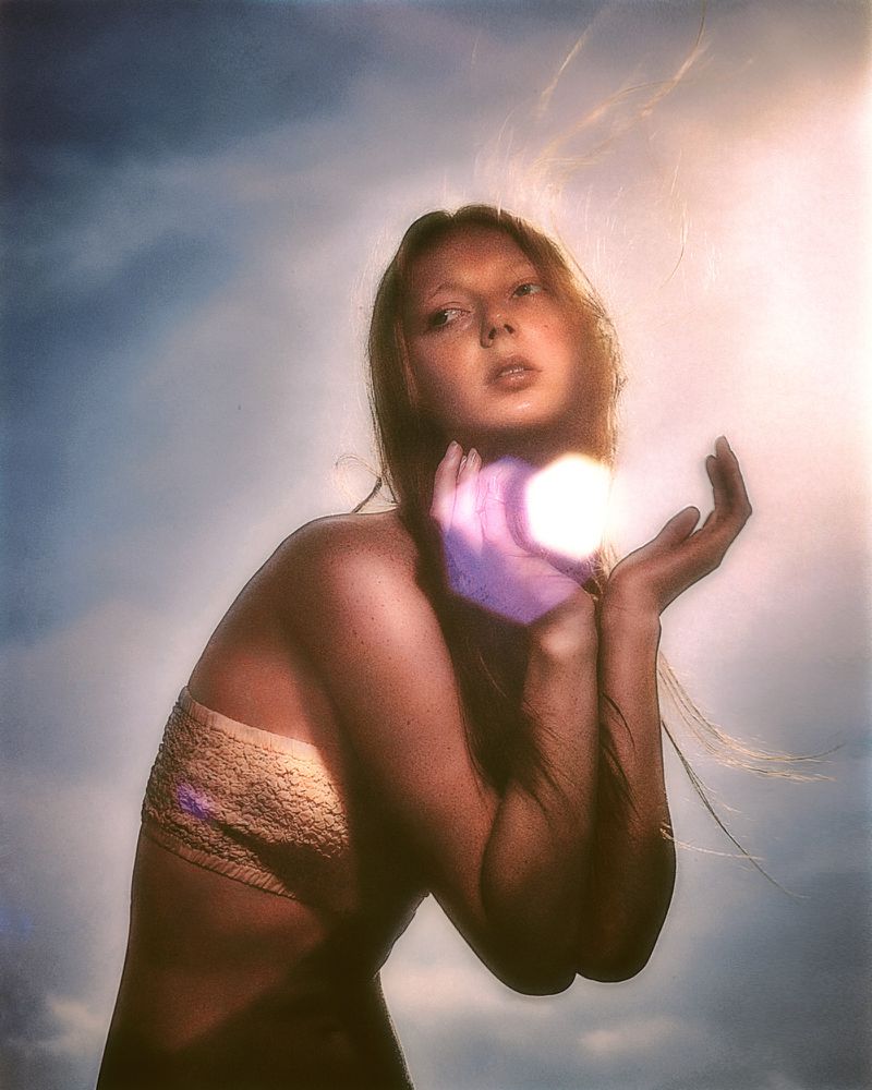 Young woman with auburn hair holds glowing orb in cupped hands against a cloudy sky, looking up with a neutral facial expression.