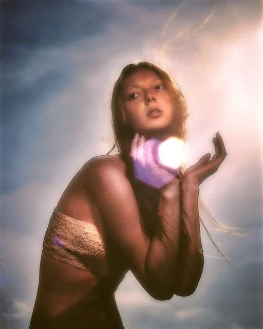 Young woman with auburn hair holds glowing orb in cupped hands against a cloudy sky, looking up with a neutral facial expression.
