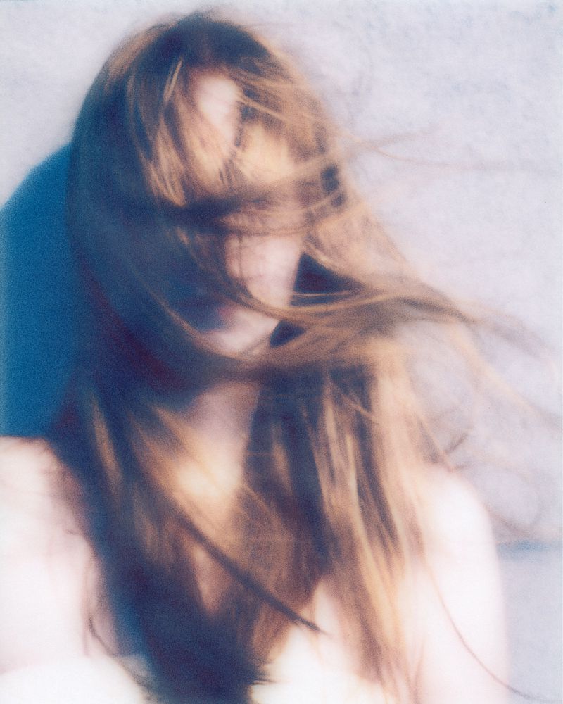 Close-up of a young adult woman with light brown hair blowing in the wind, obscuring her face, set against a neutral background.