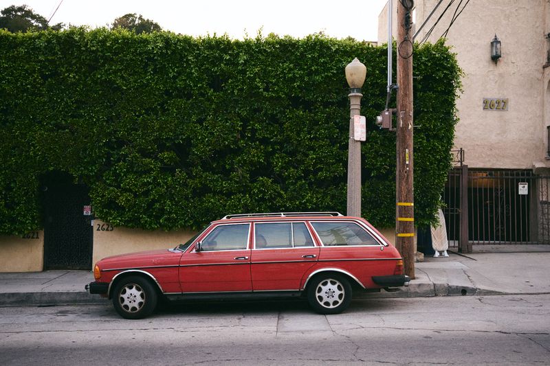 Red station wagon parked on a city street with a tall green hedge and a building in the background, providing a vintage urban scene.