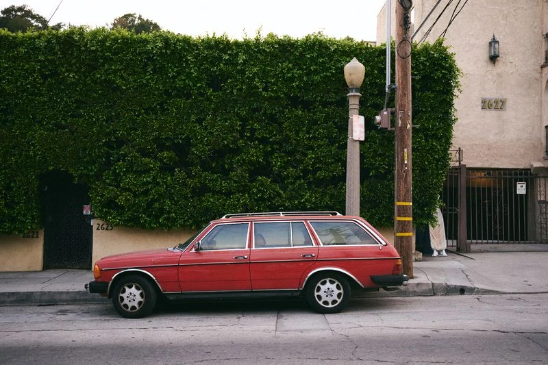 Red station wagon parked on a city street with a tall green hedge and a building in the background, providing a vintage urban scene.