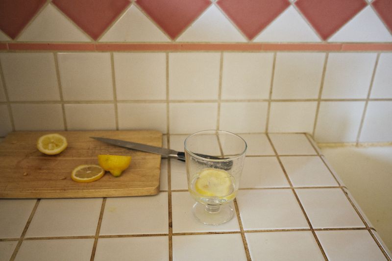 Still life of sliced lemons and a clear glass on a tiled kitchen counter with a knife and wood cutting board.