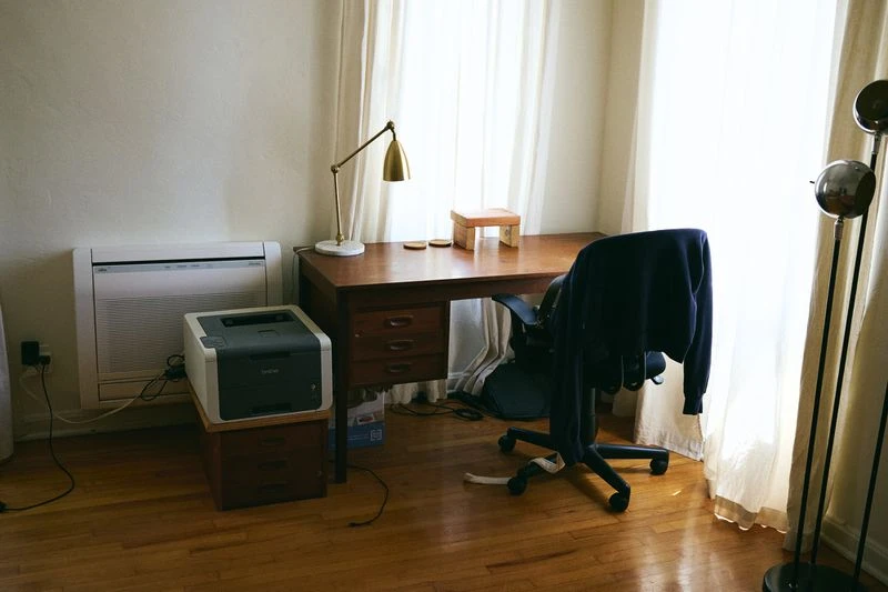 Home office setup featuring a wooden desk, chair, desk lamp, printer, air conditioner, and hardwood floors against a backdrop of white walls.