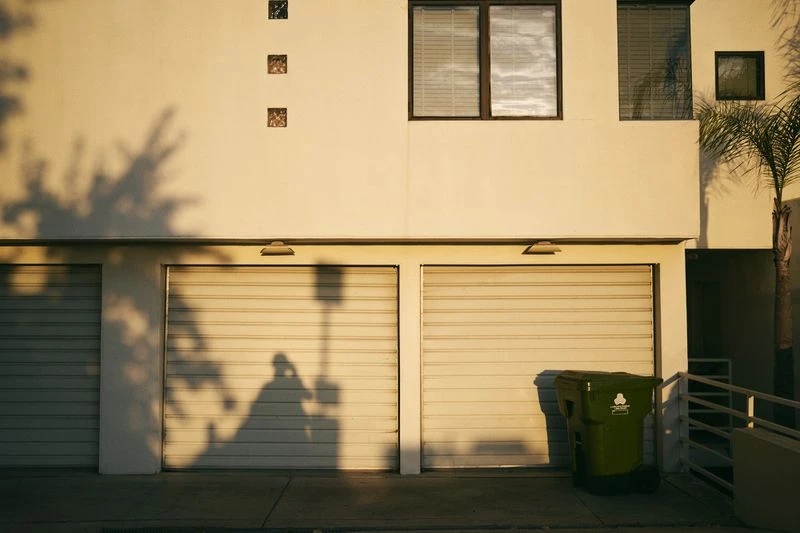 Exterior of a building with a row of garage doors, windows, and a trash can casting a shadow on a sunny day.