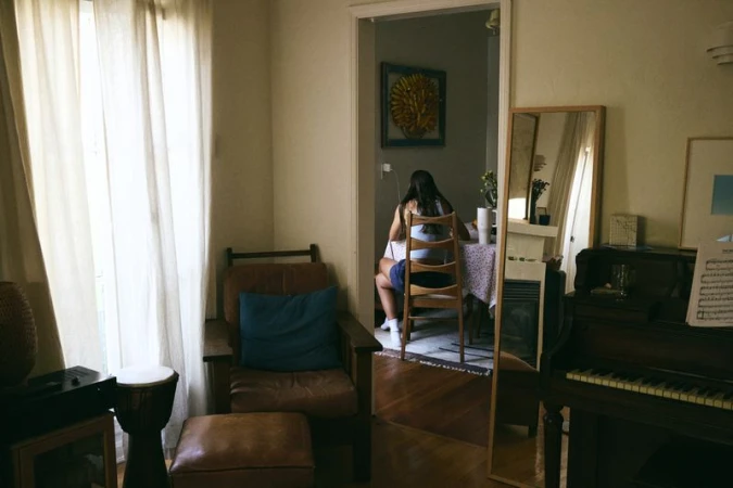 Indoor scene features a woman seated at a table, with a piano and vintage-style furnishings, bathed in soft, natural light coming through a window.