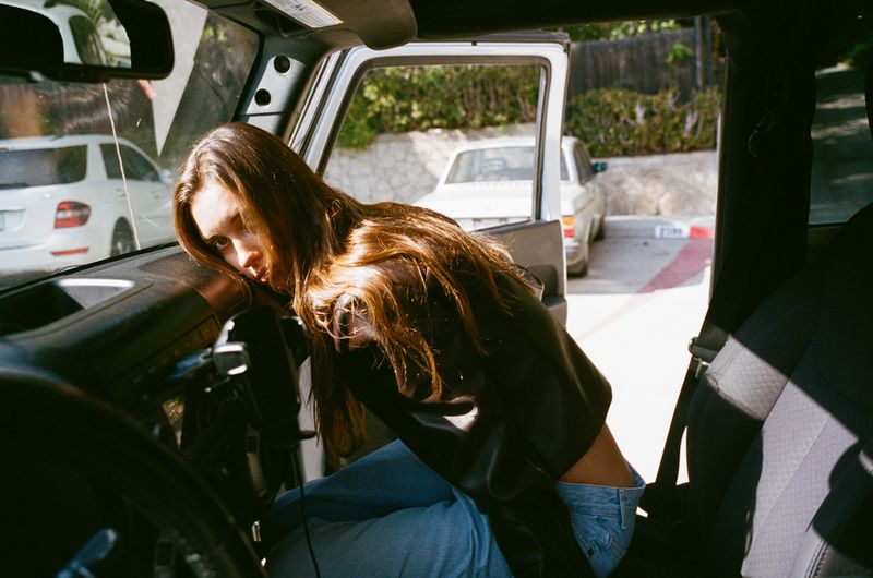 Brunette woman with long hair rests on the interior of an open Jeep Wrangler, while parked outside with other cars in the background.