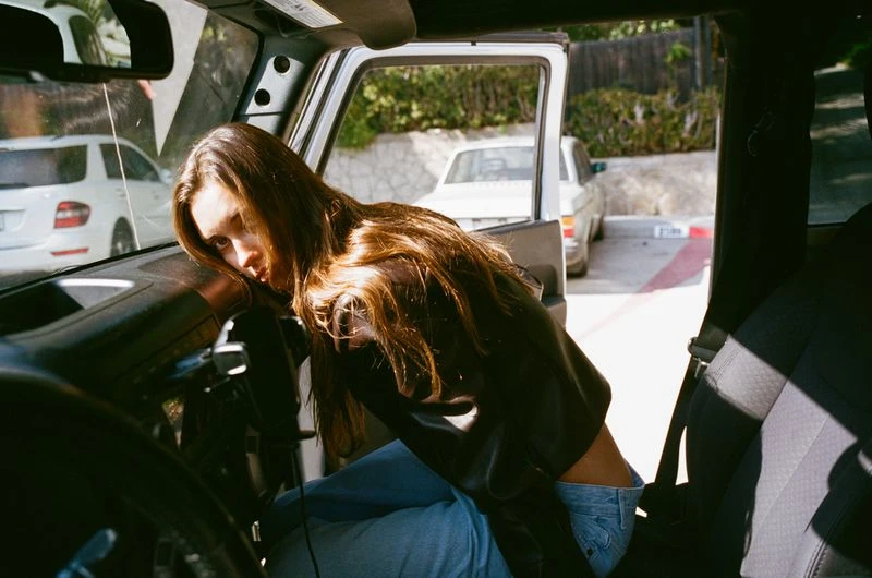 Brunette woman with long hair rests on the interior of an open Jeep Wrangler, while parked outside with other cars in the background.