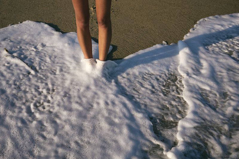 Low angle shot of a woman's legs standing in frothy ocean water at a beach, with a sandy shore visible in the background.