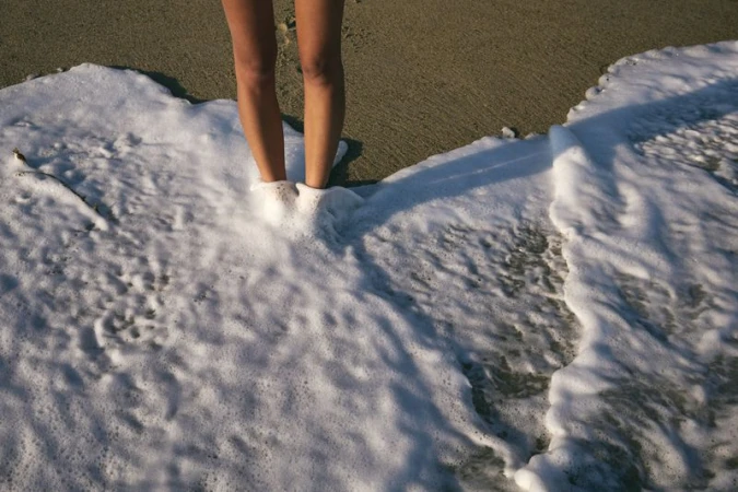 Low angle shot of a woman's legs standing in frothy ocean water at a beach, with a sandy shore visible in the background.