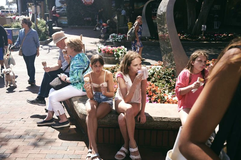 People of varying ages enjoy ice cream cones while sitting on a stone bench on a sunny day with trees and plants in the background.