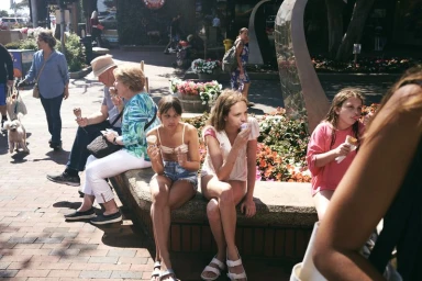 People walking and eating ice cream outdoors in a sunny public square with brick paving and flower planters