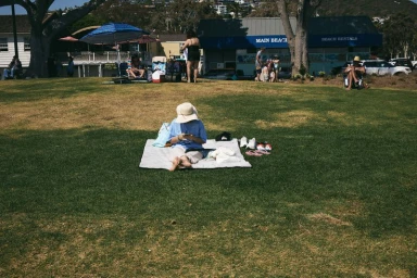 Person seated on a blanket on a grassy park lawn under bright daylight with beach buildings in the background.