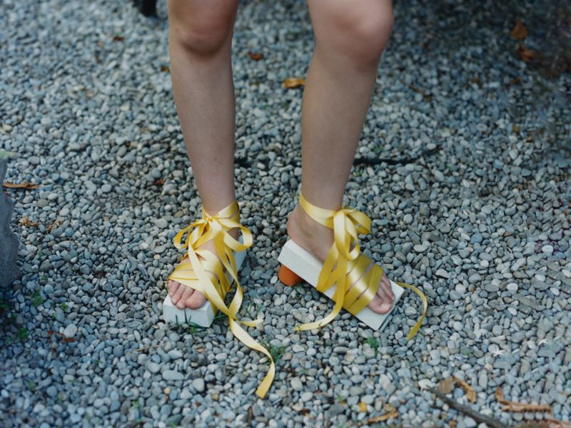 Woman stands on pebble ground wearing open-toe platform sandals with yellow ribbon ankle ties, showcasing a unique fashion statement.