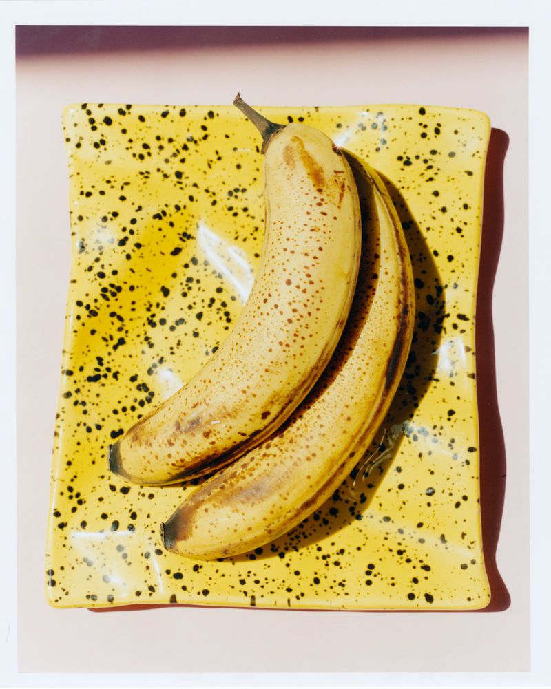 Overripe bananas on a yellow speckled square plate, set against a light pink background in a brightly lit studio shot.