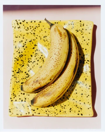 Overripe bananas on a yellow speckled square plate, set against a light pink background in a brightly lit studio shot.