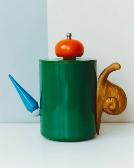 Whimsical green ceramic teapot with a mushroom lid, a blue spout, and a snail-shaped handle sits on a white surface against a blue and white backdrop.