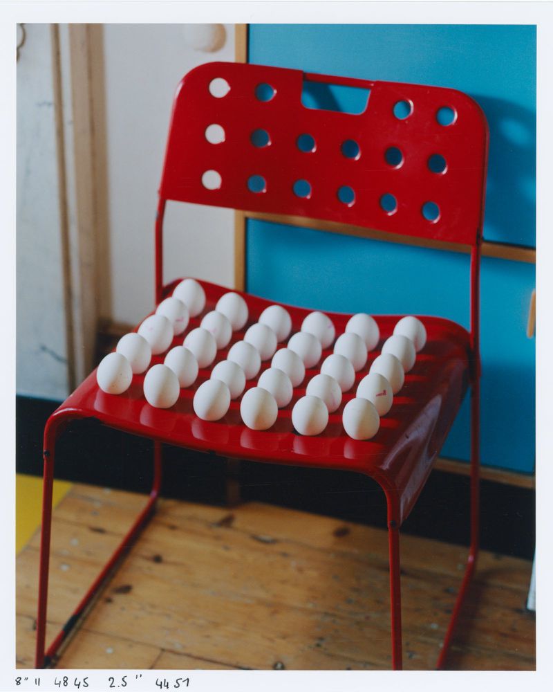 Eye-level shot of a red metal chair indoors. The chair seat is covered with rows of white chicken eggs against a studio backdrop.