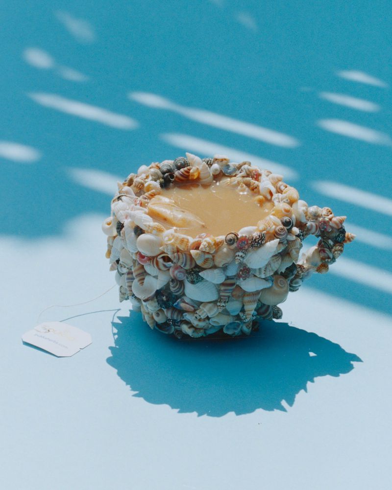 Teacup covered in seashells filled with liquid resting on a light blue surface casting shadows with white tag attached to handle.