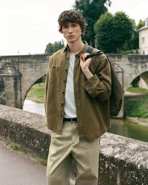 A male model stands on a stone bridge wearing a brown button-down shirt, white t-shirt, and khaki pants with a shoulder bag.