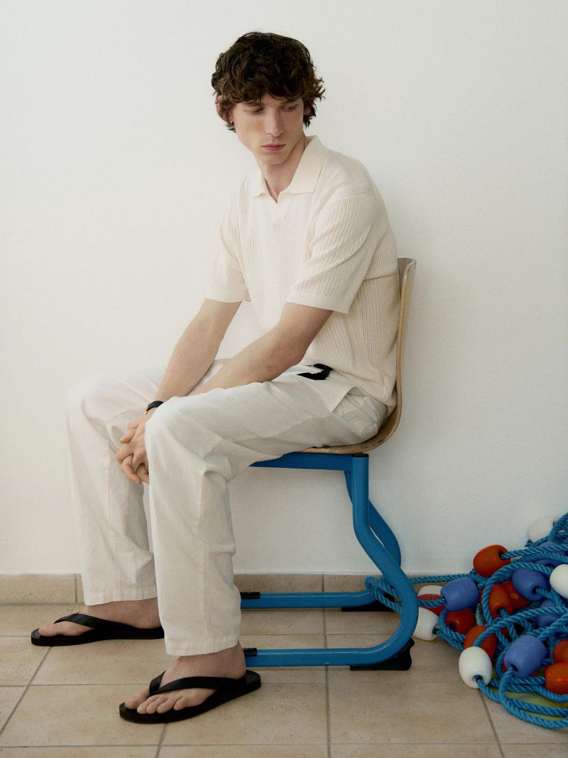 Adult male fashion model sits on blue chair in cream shirt, light pants, and black flip-flops in a neutral indoor setting near ropes.