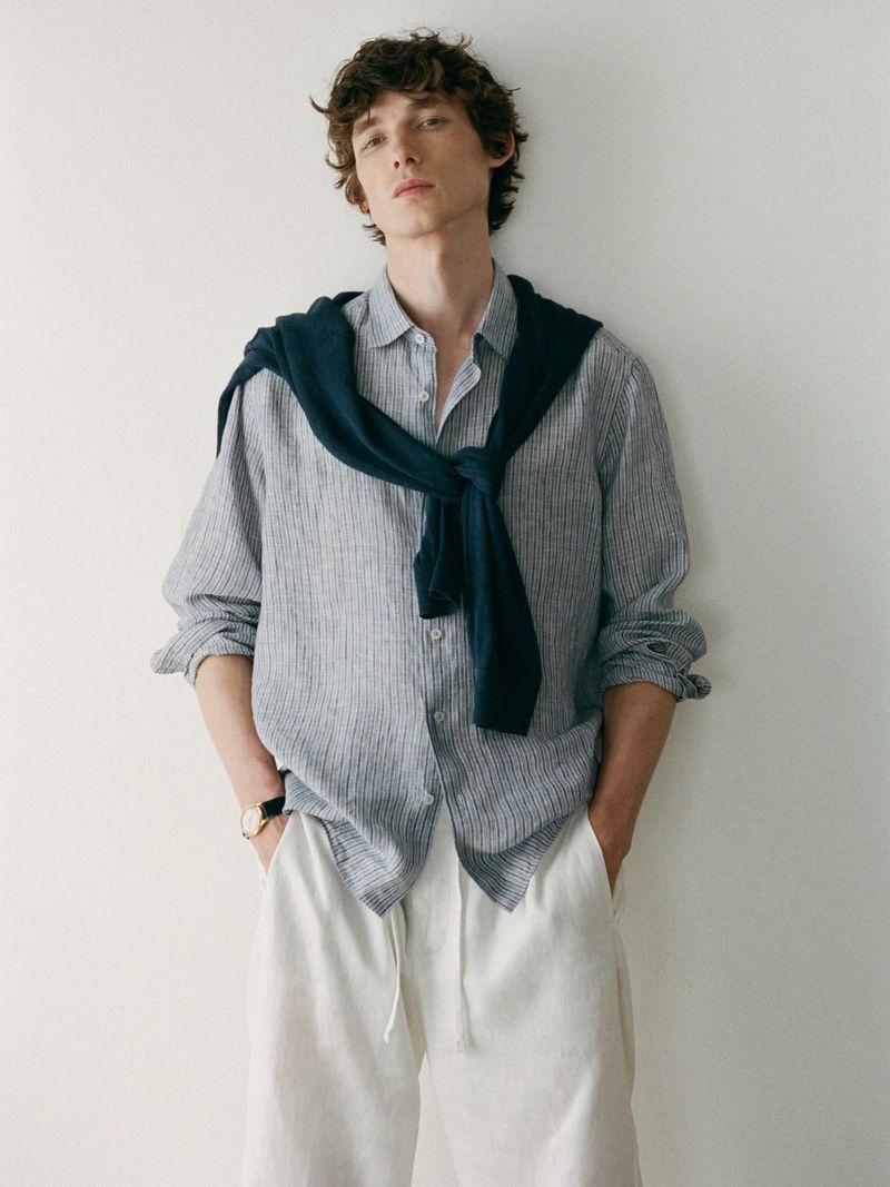 A young adult man is posing indoors wearing a striped shirt, white pants, and a navy sweater draped over his shoulders, looking at the camera.