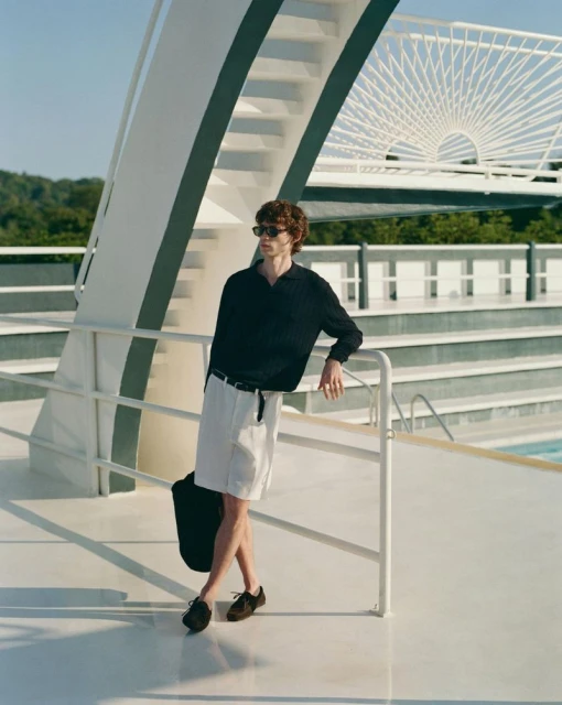 Man in black shirt and white shorts leans on a white railing near a pool on a sunny day, styled in casual fashion.