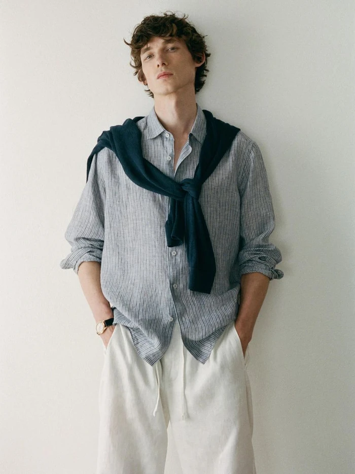 Male model wearing a striped shirt with a dark sweater tied over his shoulders and white trousers against white