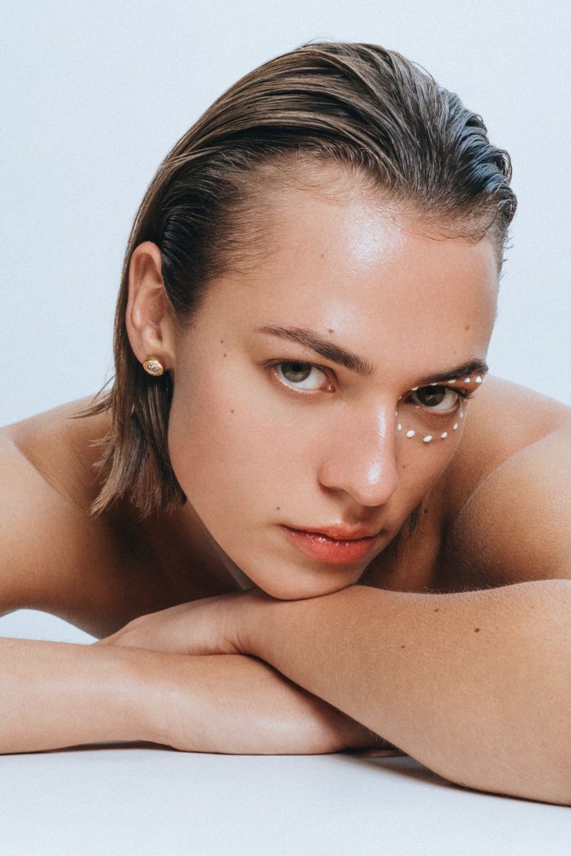Close-up portrait of a fair-skinned woman with wet, dark blonde hair, light eyes, and small pearls applied under her eye.