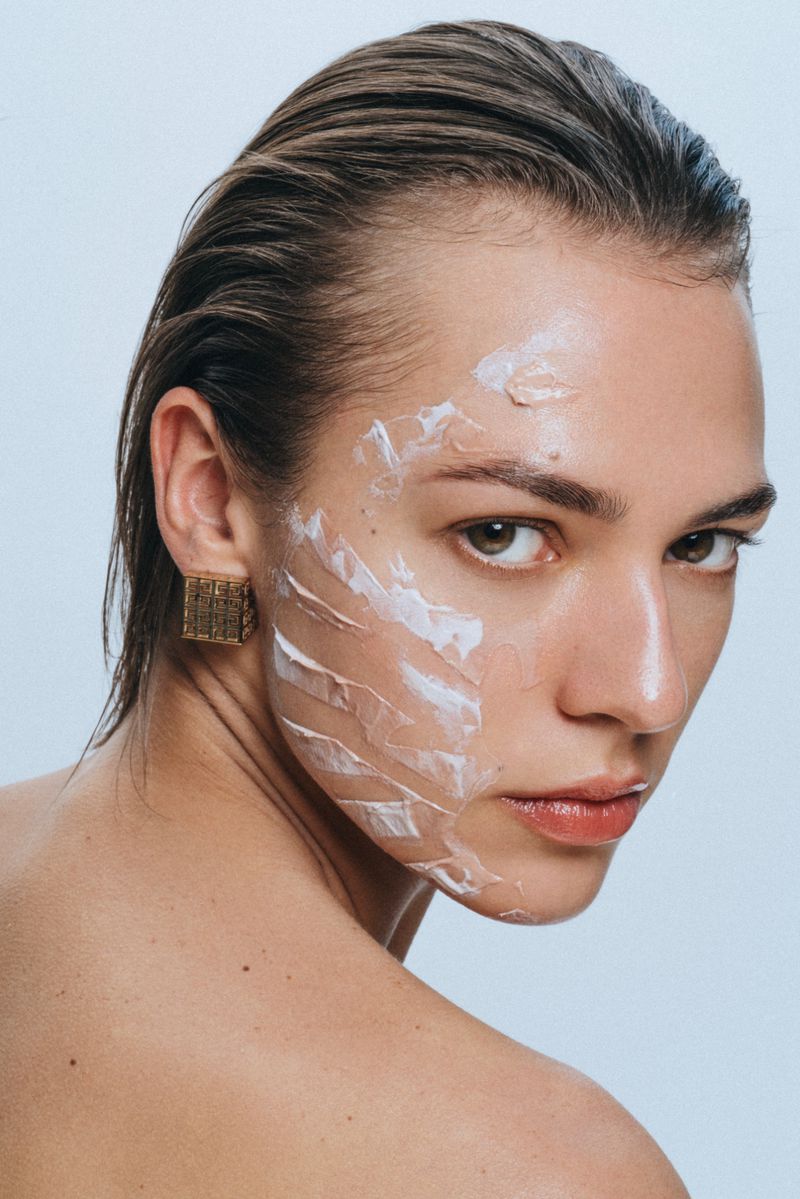 Close-up of a young adult female with an asymmetrical haircut, wearing an earring, and white cosmetic cream on half of her face.