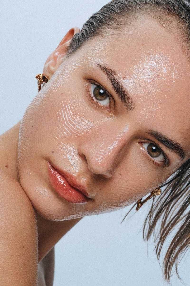 Close-up portrait of a young adult woman with wet-look hair and glossy skin, wearing gold earrings, against a light blue background.