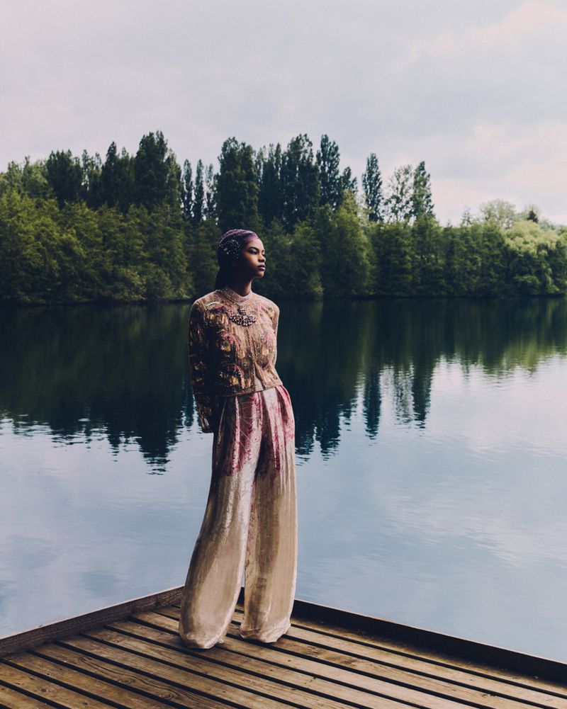 Woman in an embellished top and wide leg pants stands on a wooden dock by a lake with trees in the background on an overcast day.
