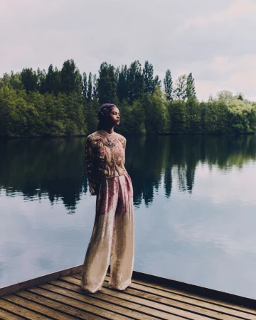 Woman in an embellished top and wide leg pants stands on a wooden dock by a lake with trees in the background on an overcast day.