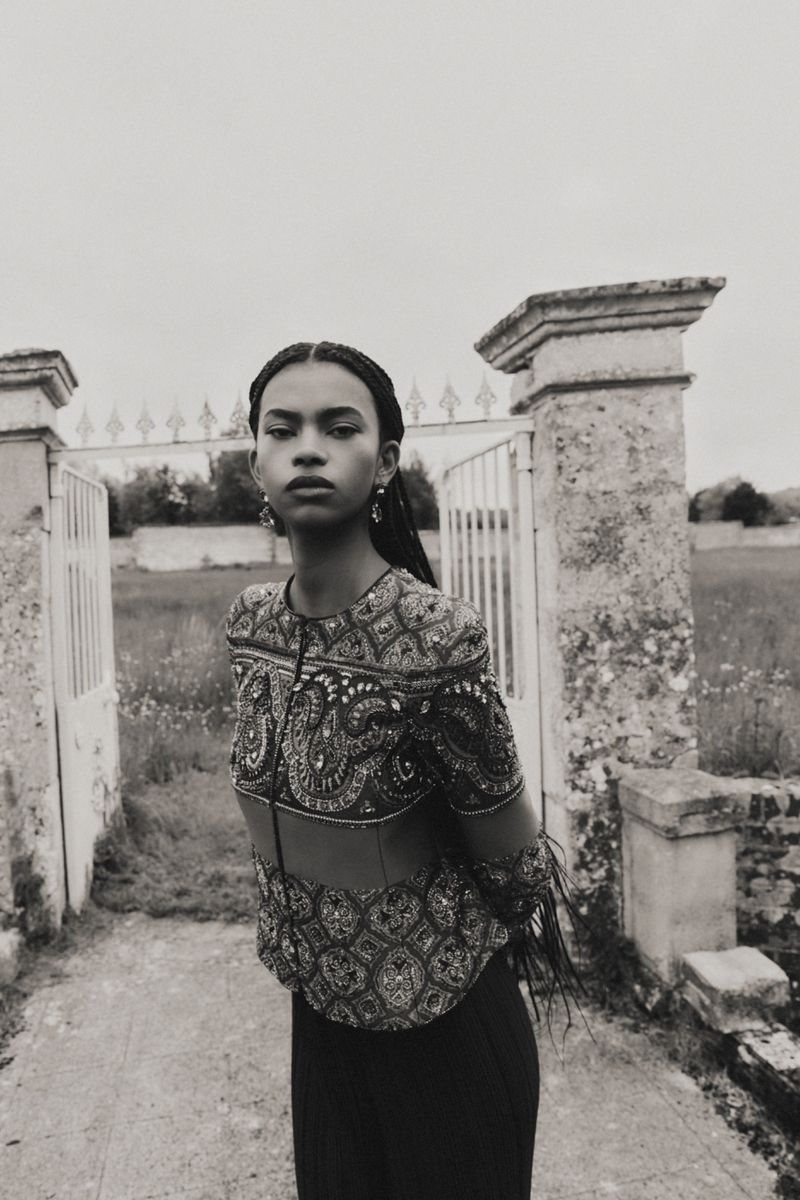 Portrait of a woman with braided hair wearing an ornate top, standing in front of an old gate in a rural setting, grayscale image.