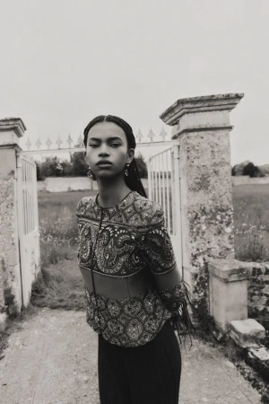 Portrait of a woman with braided hair wearing an ornate top, standing in front of an old gate in a rural setting, grayscale image.